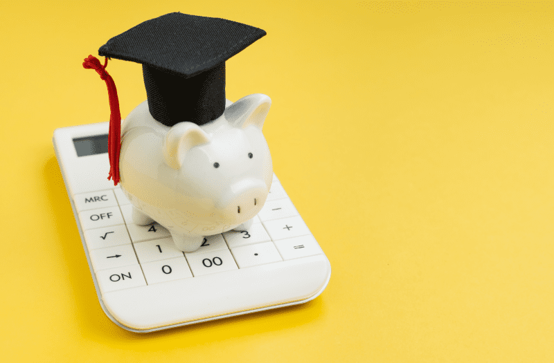 Alcancía de cerdito con un birrete de graduado, arriba de una calculadora realizando en análisis de las becas universitarias